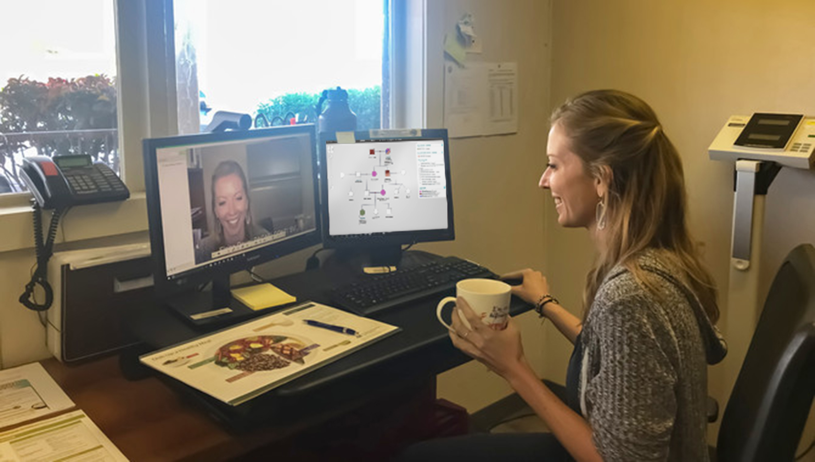A woman sits at her home office desk holding a cup of tea. She smiles and holds a mouse while looking at one of two monitors on her desk. One monitor shows she's on a video call, the other has an open PhenoTips pedigree.