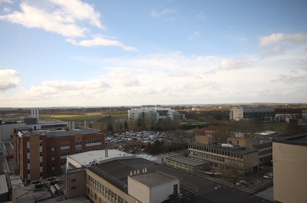 A picture of Addenbrooke’s Hospital as seen from the window of a tall building nearby. The sky is blue and clear with a few wispy clouds, and a green field speckled with cloud shadows is visible behind the white hospital building and full parking lot.