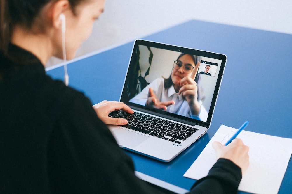 A young female genetic counselor is on a video call on her laptop while writing notes on a piece of paper. The patient in the video call appears to be explaining something.